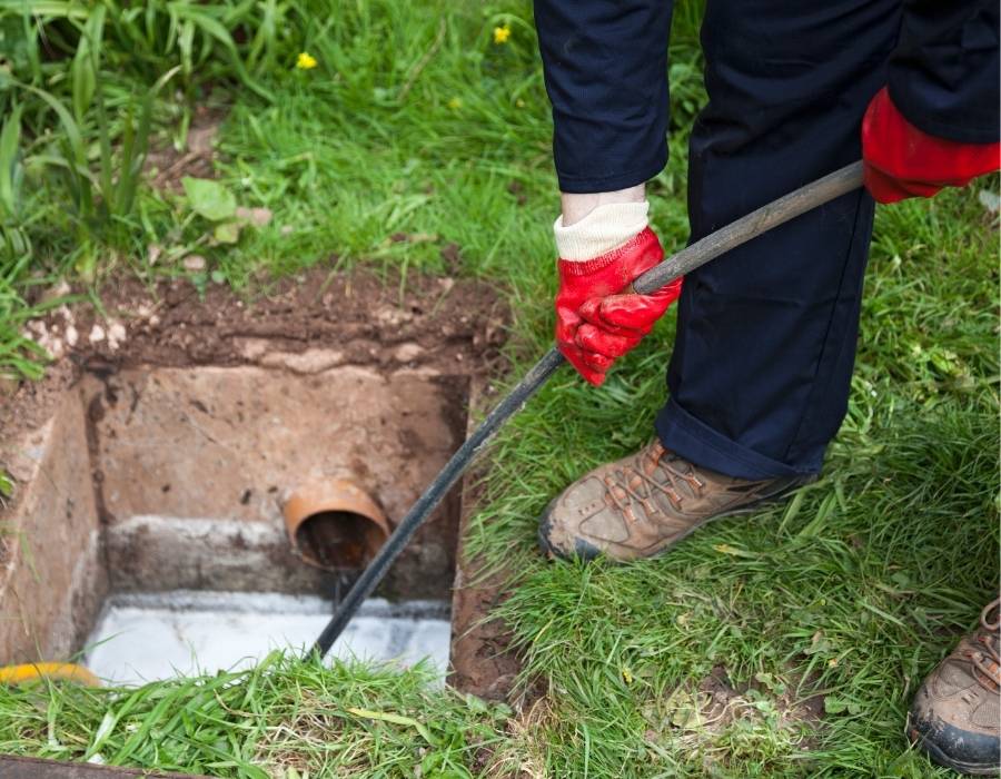 Plumber performing routine drain cleaning with manual rod to clear outdoor sewer pipe in residential yard